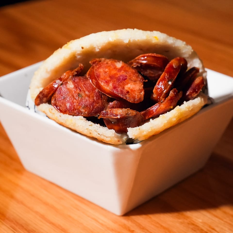 A Colombian chorizo arepa, filled with sliced sausage, served in a white dish on a wooden table.