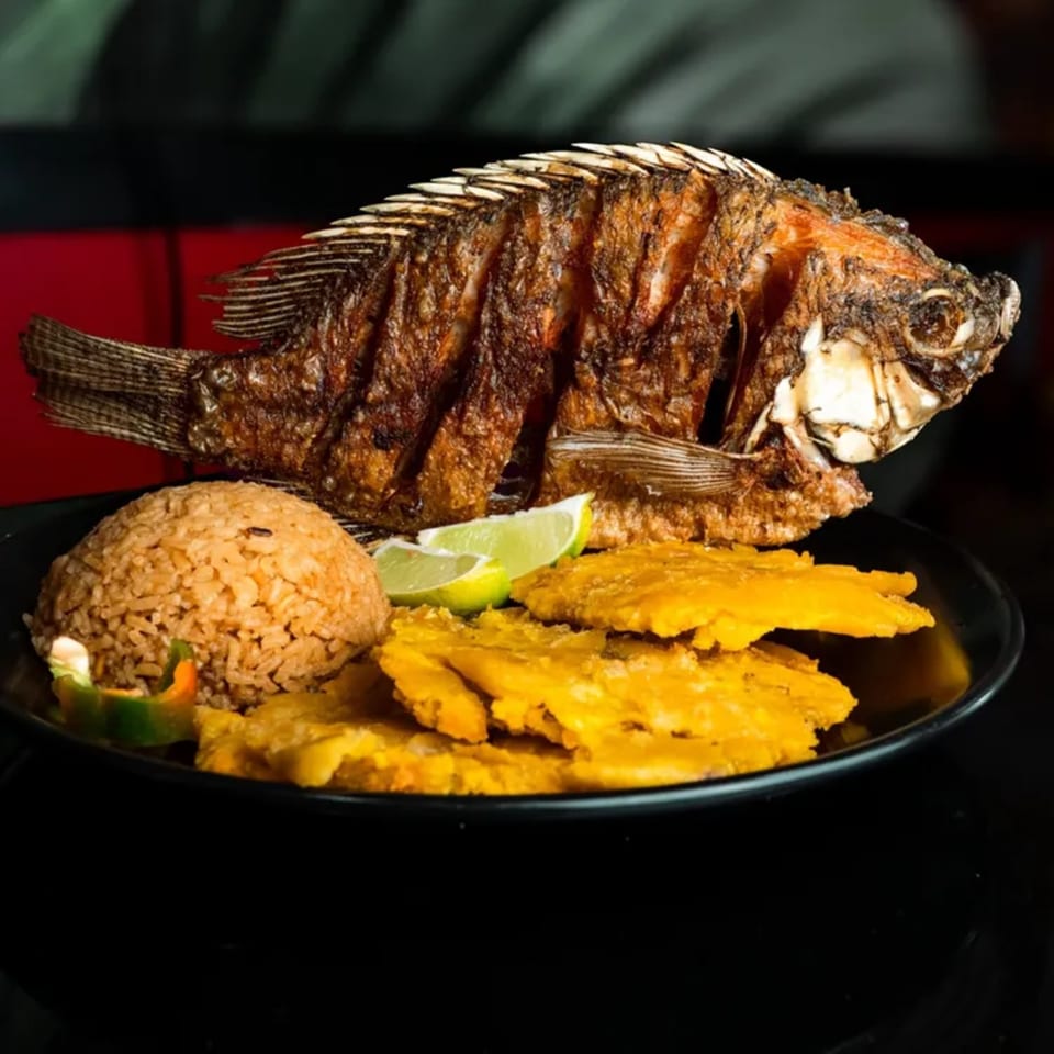 A whole fried fish with rice, lime wedges, and patacones on a black plate.