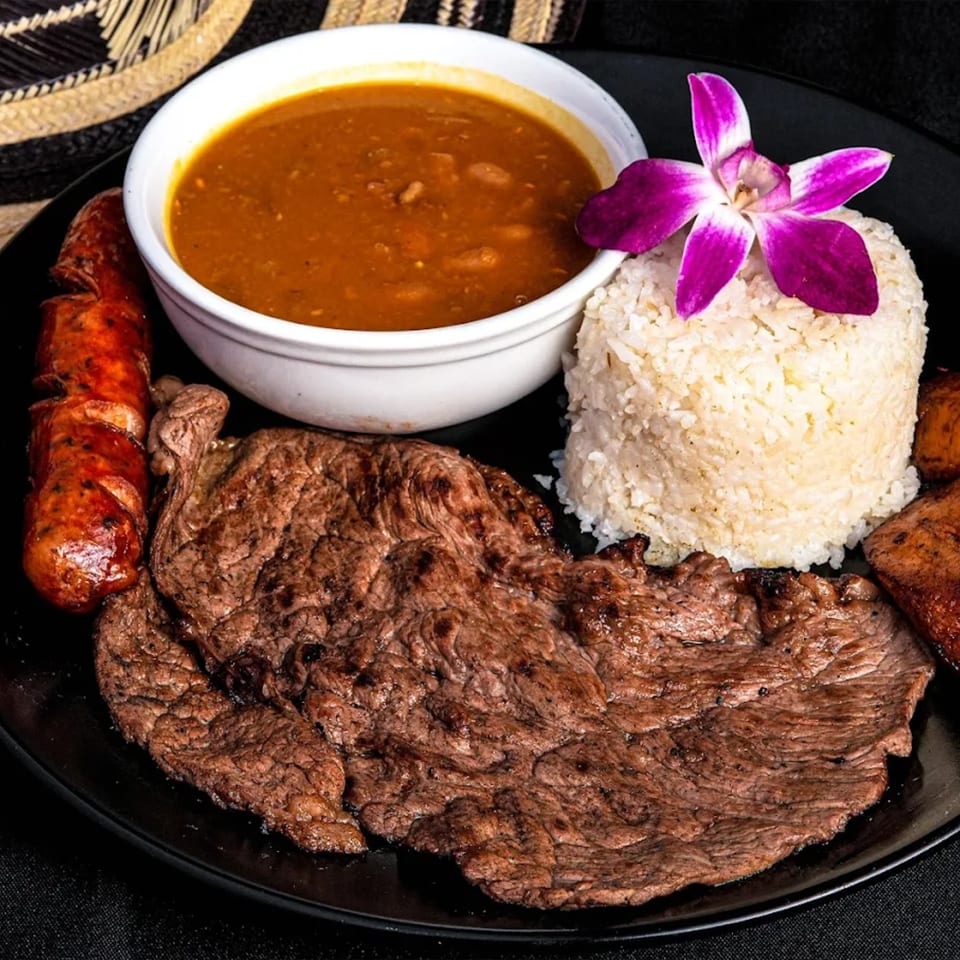A Colombian Bandeja Paisa with steak, sausage, rice, beans, and plantains, garnished with an orchid.